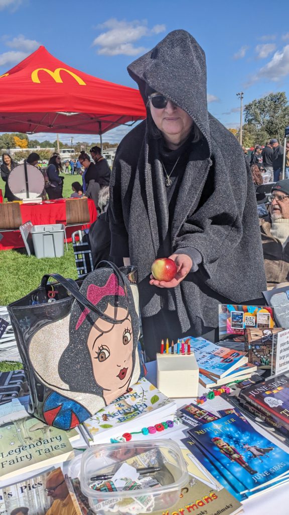 woman in black cape holds an apple next to a bag depicting Snow White's face, over a table of books