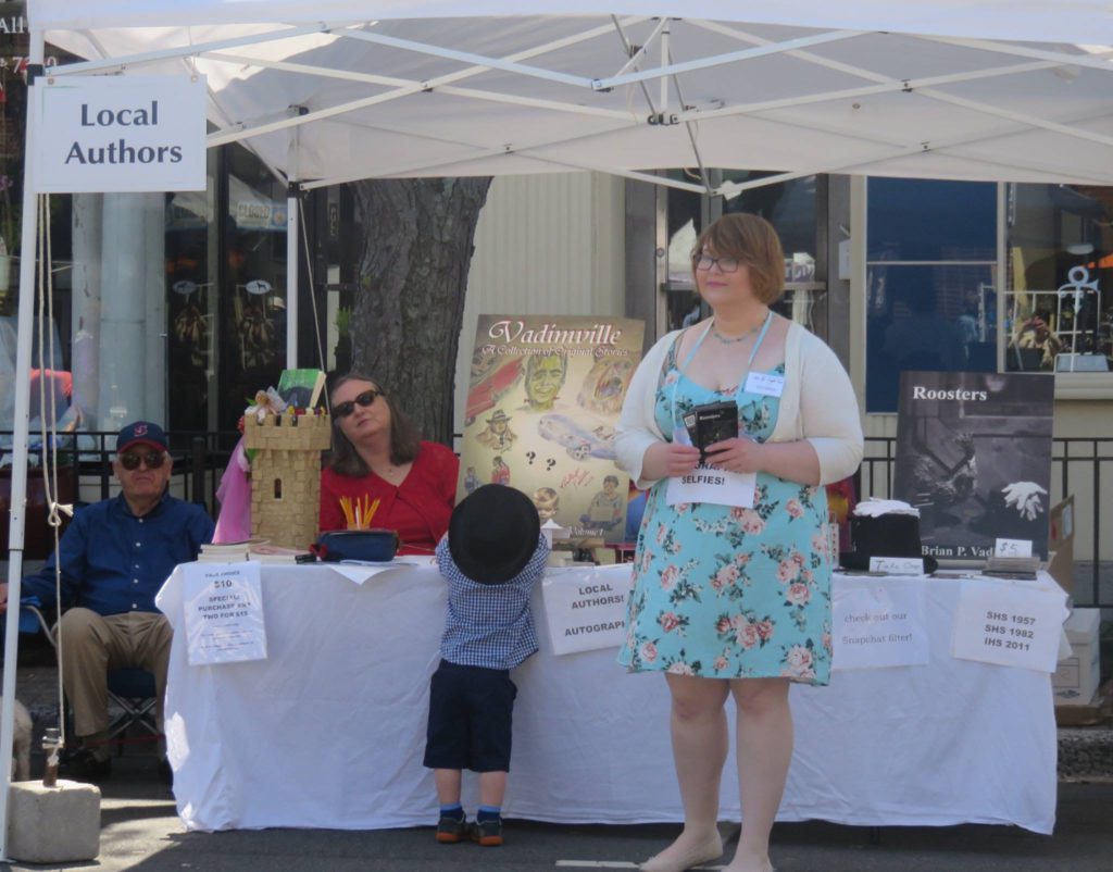 Table under a white tent labeled text "Local Authors", white tablecloth, books and posters, a woman in red with sunglasses, a young woman in flowered dress holding a book, and a little boy in a hat reaching for the table