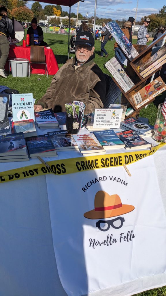 Man in cap and jacket sits at a table with books, "Crime Scene" caution tape around it