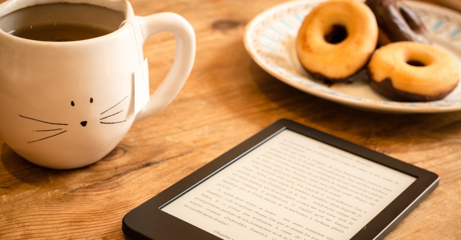 cat mug with tea on wooden table next to ebook reader and plate of donuts