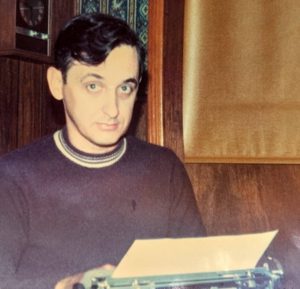 young man, Richard Vadim, with black hair wearing a crew neck sweater looks up from his work at a typewritere